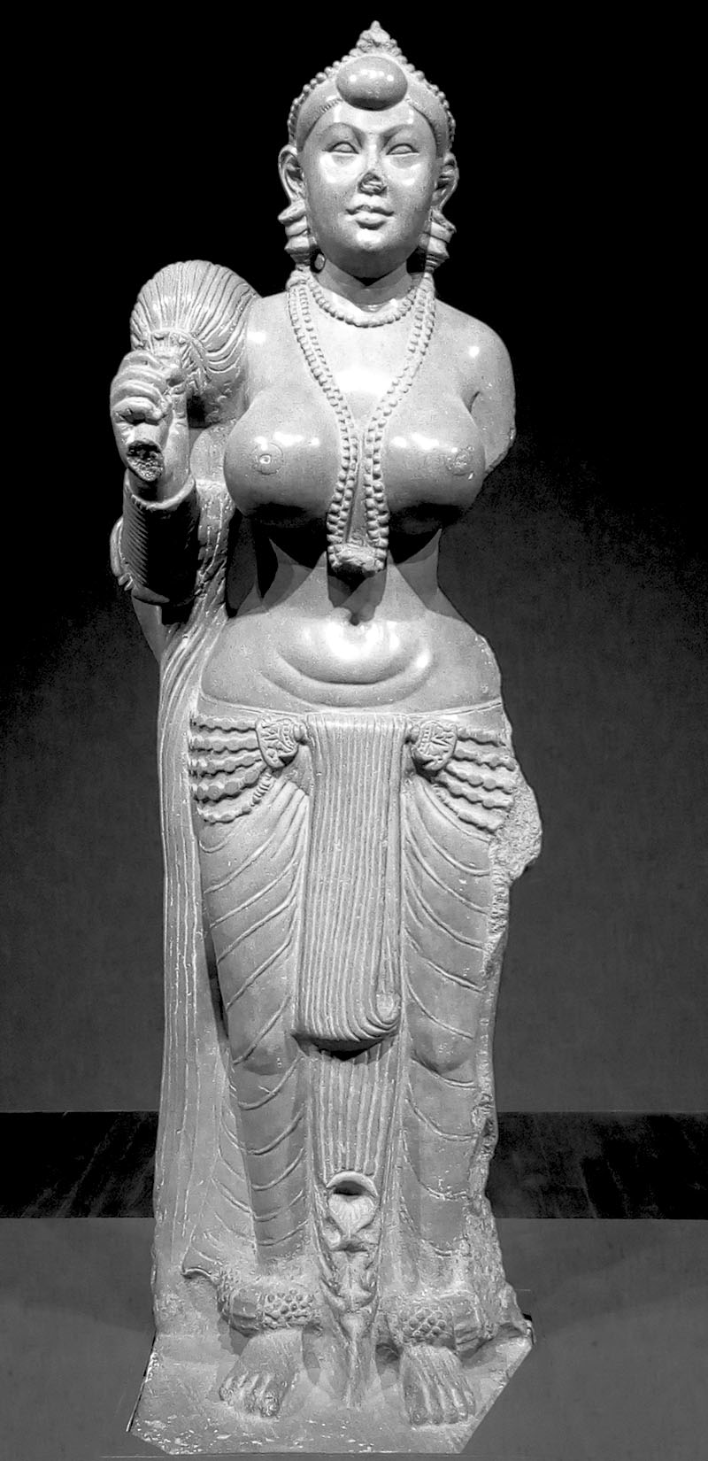 A sandstone sculpture of a voluptuous and bejewelled woman, holding a mace in her right hand and wearing a waistcloth.