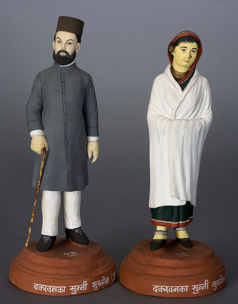 Photograph of pair of clay figurines, man dressed in grey achkan and woman in sari covered by white shawl