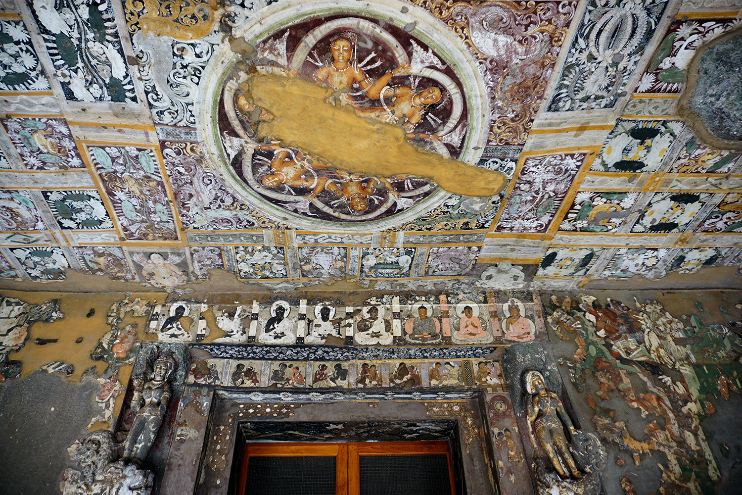 Photograph of Ajanta Cave 17 interior with multicoloured ceiling and wall murals showing Buddhas, other figures and various patterns
