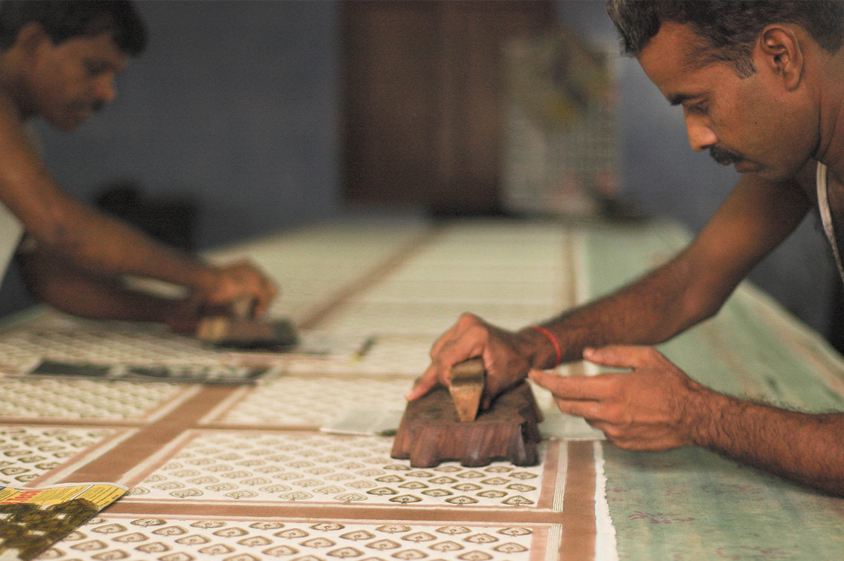 Photograph of two artisans using wooden blocks to print motifs on a large piece of cloth laid flat on a table