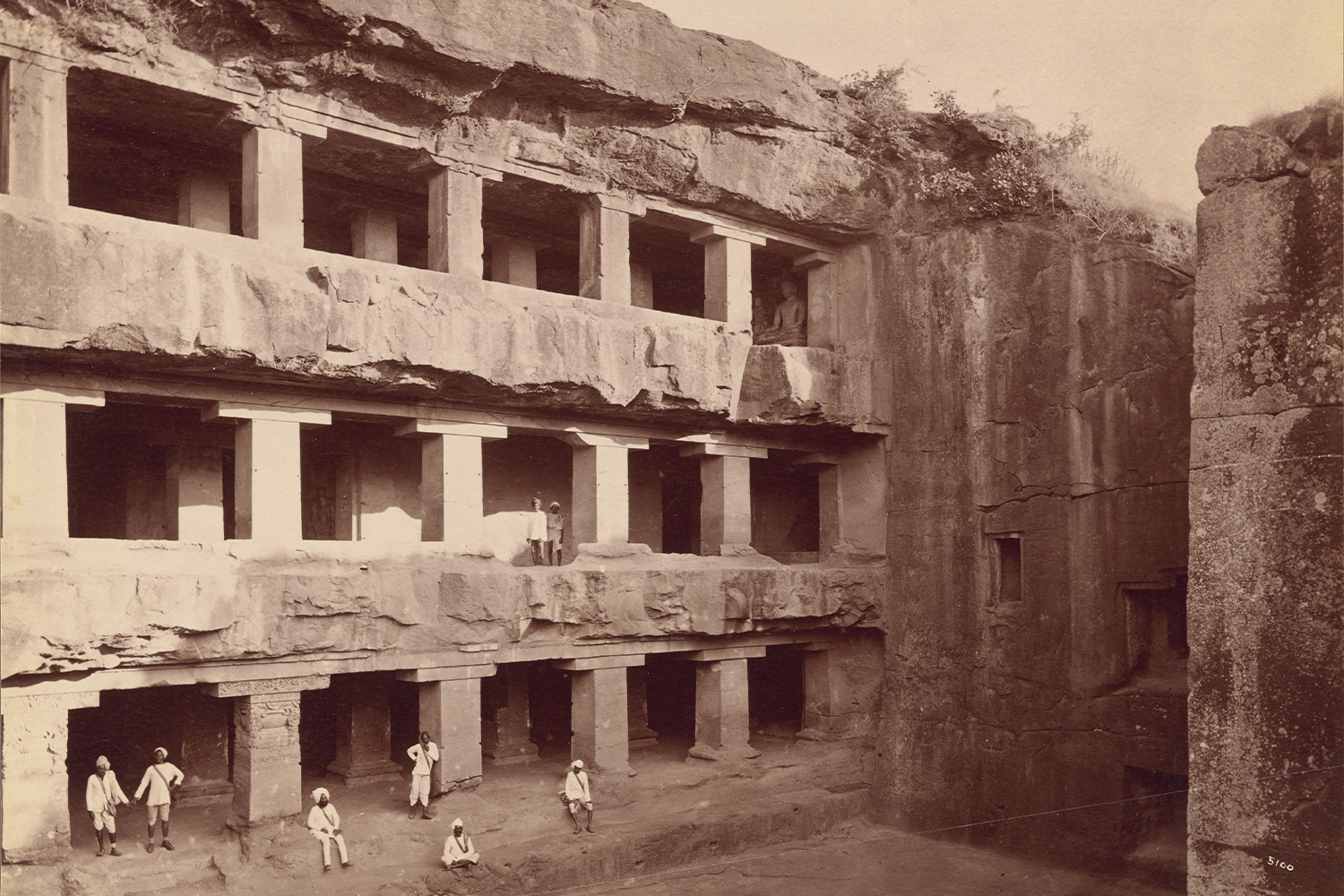 Monochrome albumen print by Lala Deen Dayal showing the facade of the 3-storey Tin Taal Cave at Ellora, with people seen at the lower and 2nd storeys