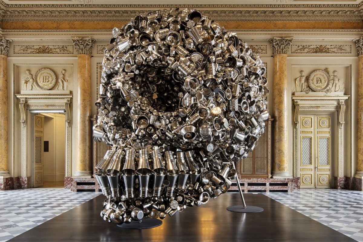 Floor-to-ceiling art installation by Subodh Gupta showing steel utensils arranged together to resemble a human skull titled Very Hungry God