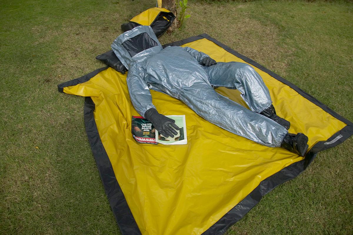 Photograph of an untitled work by Mariam Suhail showing a figure in a hazmat suit lying down on a yellow mat with a magazine