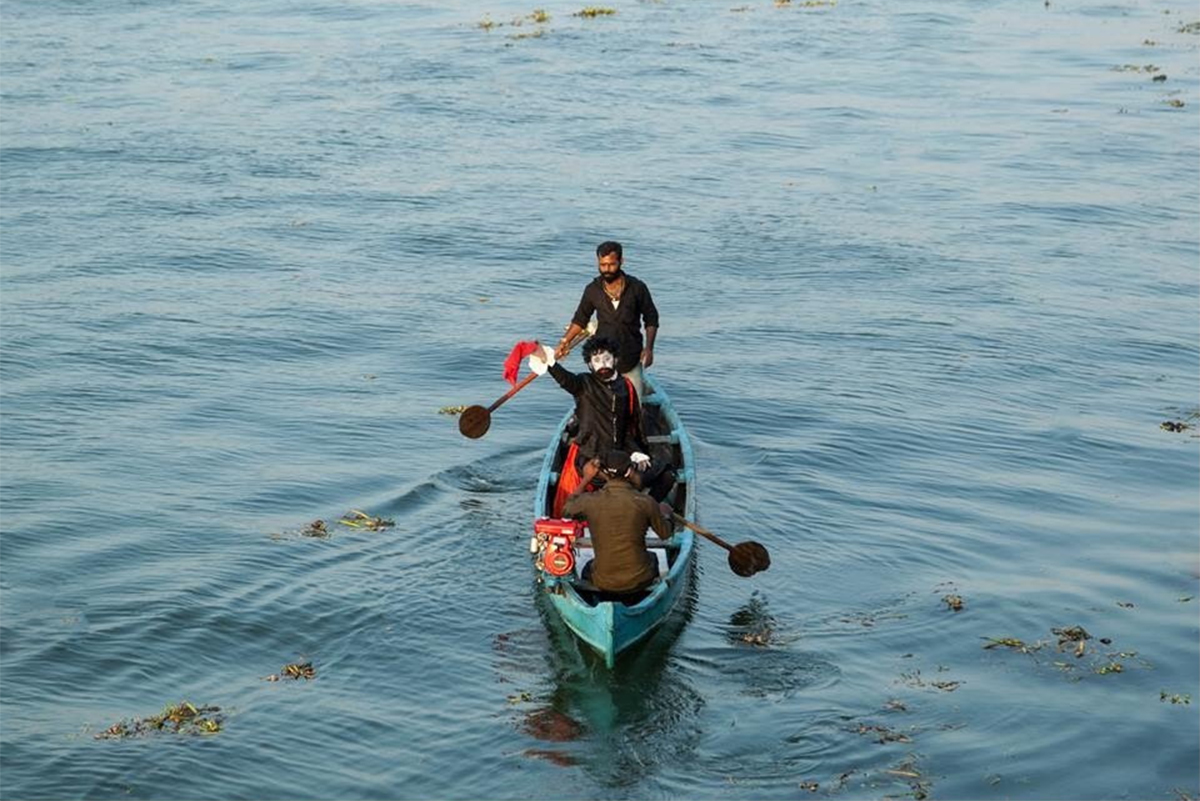 Photograph from a performance art by Nikhil Chopra called La Perle Noire (or the Black Pearl) showing a man with makeup in a boat waving at the camera as two other people row the boat