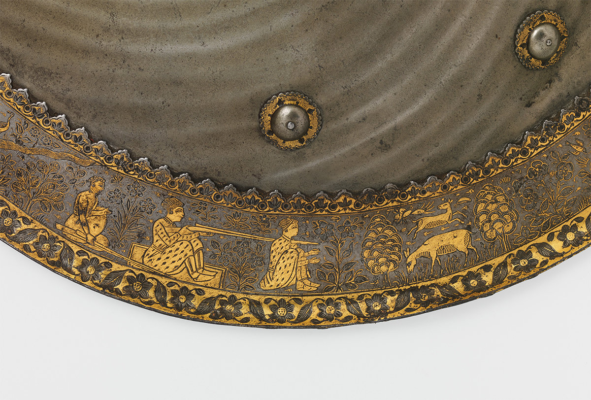 Close-up photograph of the gold overlay on the rim of a steel shield showing a hunting scene
