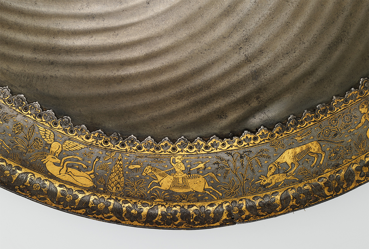 Close-up photograph of the gold overlay on the rim of a steel shield showing a hunting scene on horseback and a dog hunting a hare
