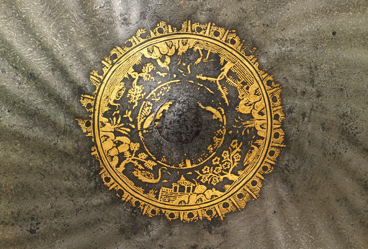 Close-up photograph of the gold overlay on the knob of a steel shield showing a landscape in a roundel
