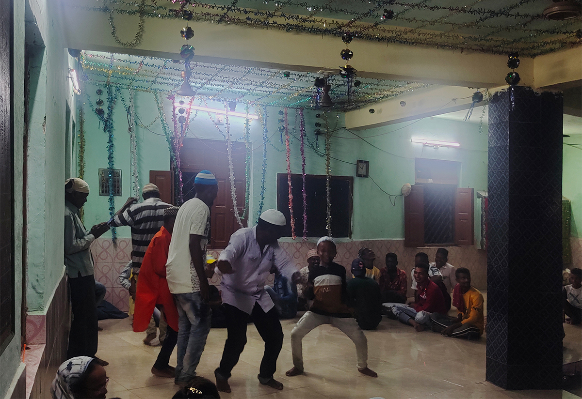 Photograph of some Siddi men and boys doing dancing dhamaal with others watching seated, at a shrine in Gujarat