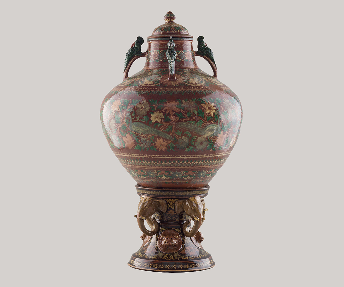 Photograph of a glazed ceramic vase with a stand and a lid, with floral designs on a red background; parrot- and elephant-shaped handles