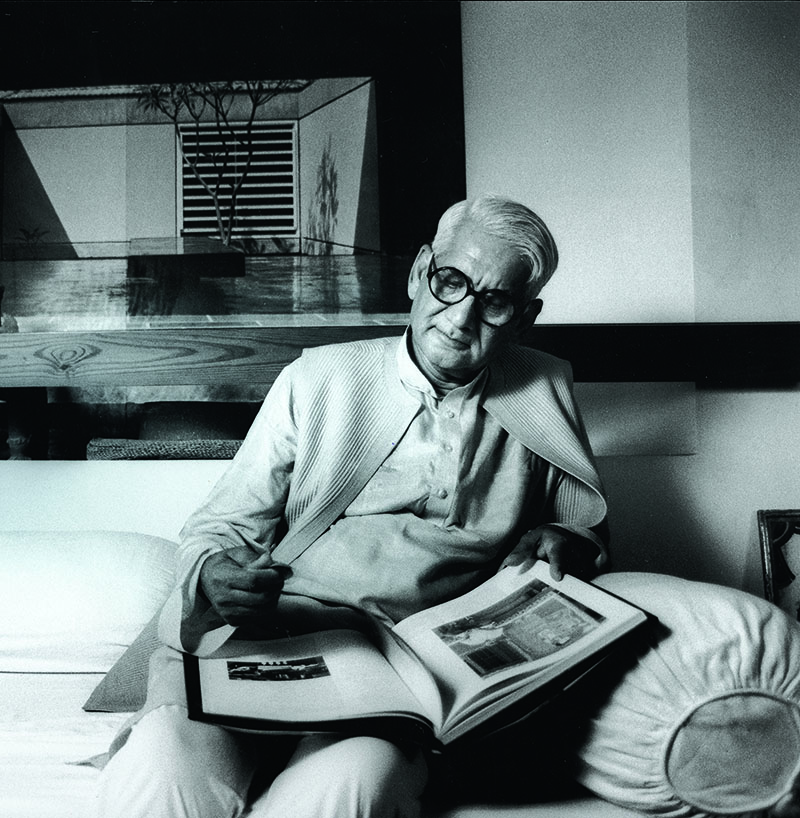 Photograph of Charles Correa seated on a couch with a book