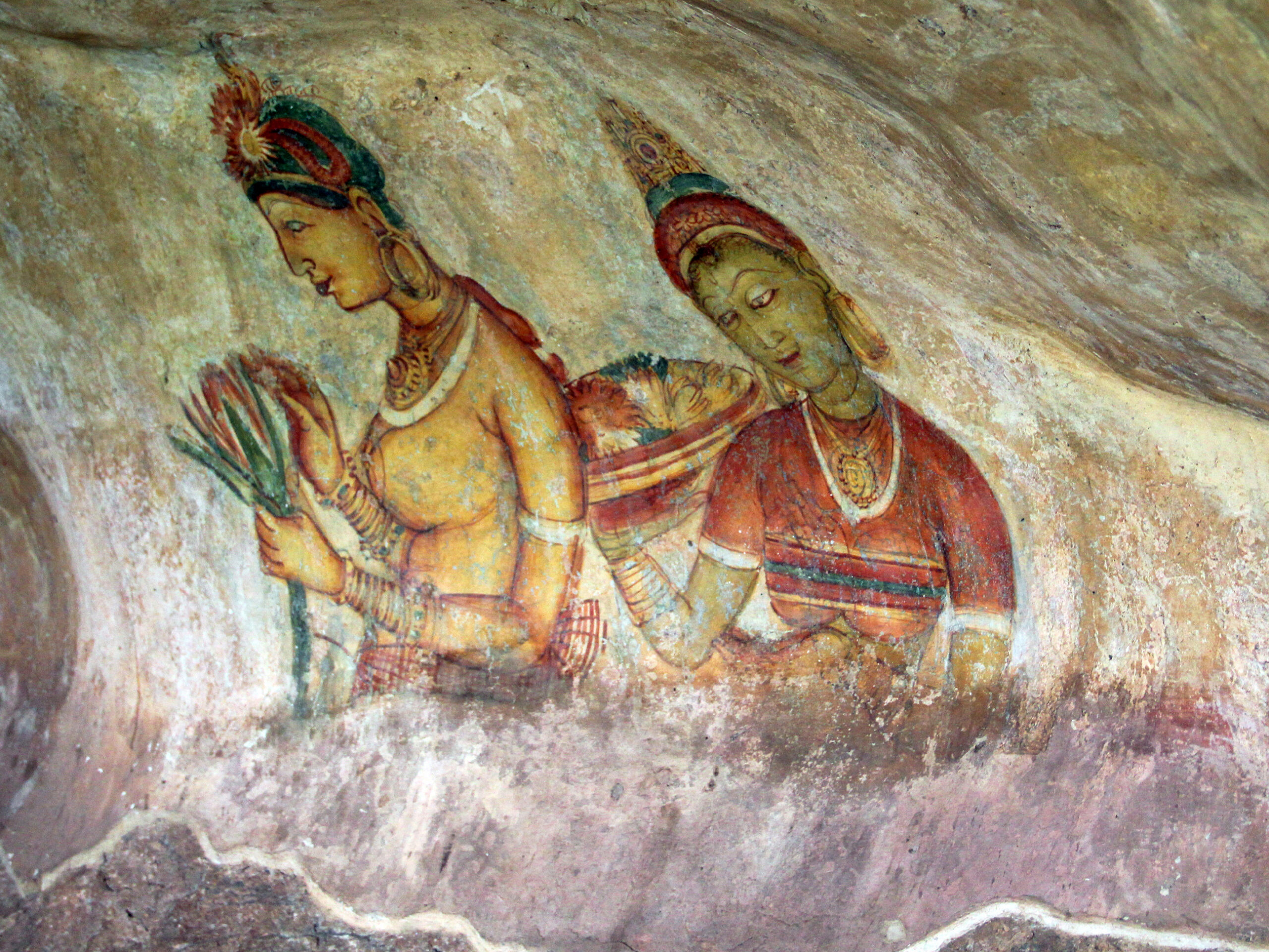 Photograph of a Sigiriya mural showing two women, both heavily bejewelled, with one holding a flower, the other holding a tray full of flowers
