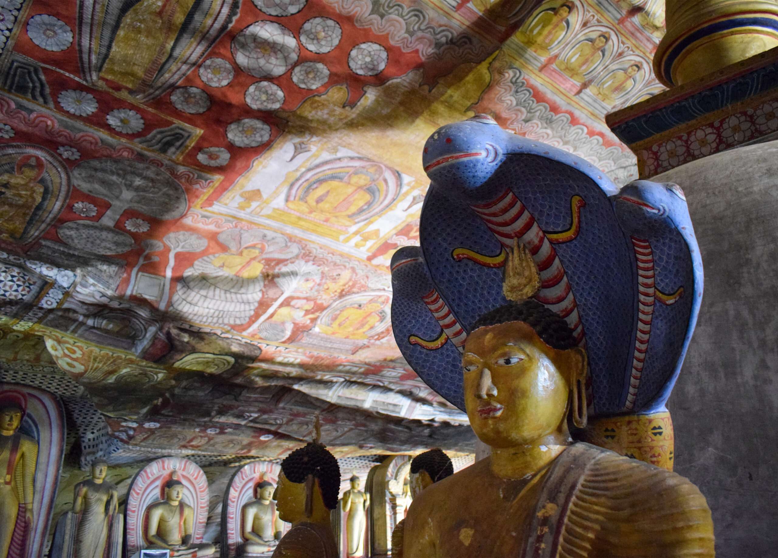 Close-up photograph from inside the Dambulla Caves of a seated Buddha with his head covered by the hood of a snake, in the background murals can be seen on the ceiling