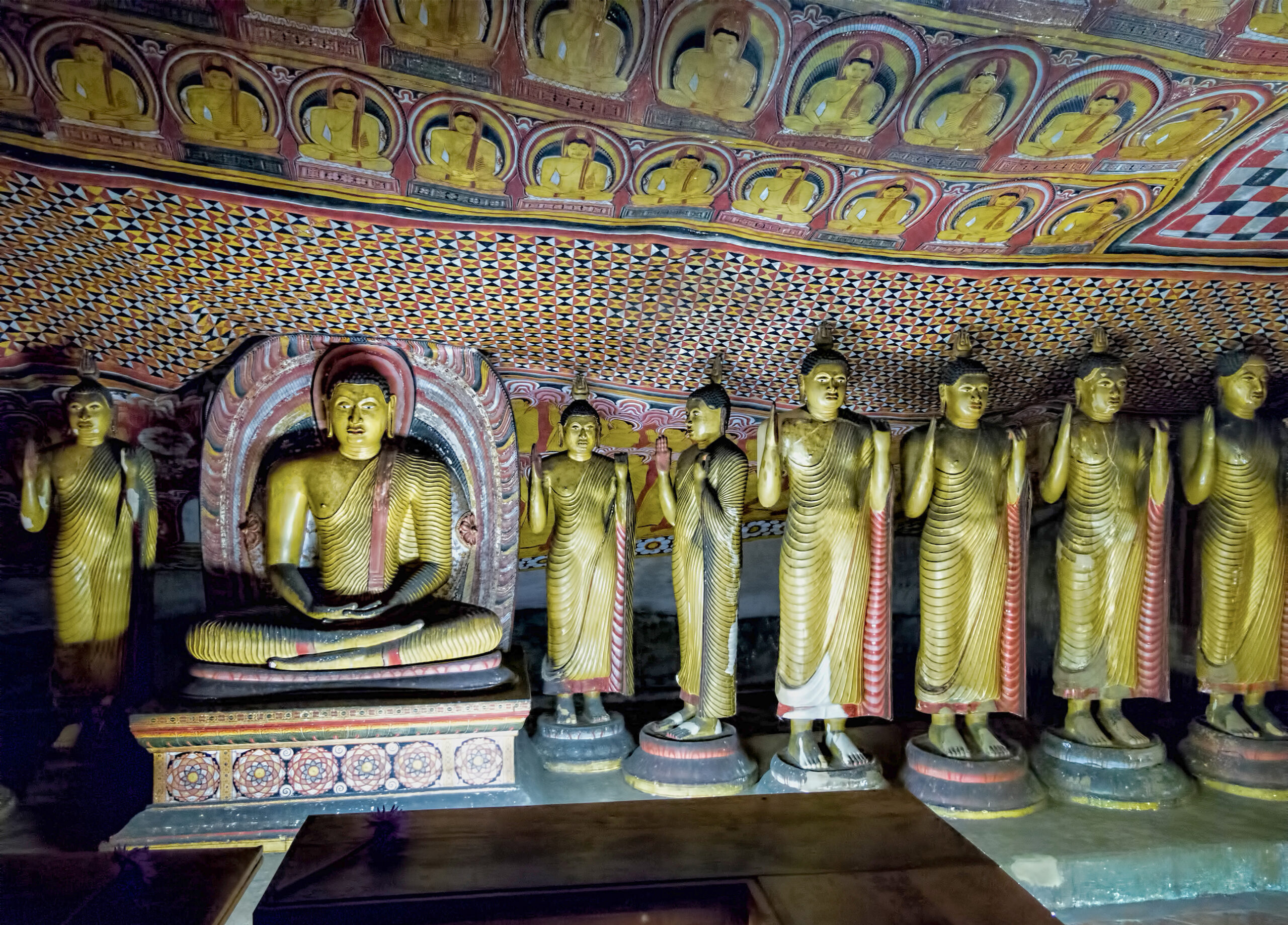 Photograph showing a seated and multiple standing Buddhas with murals on the ceiling inside the Dambulla Caves