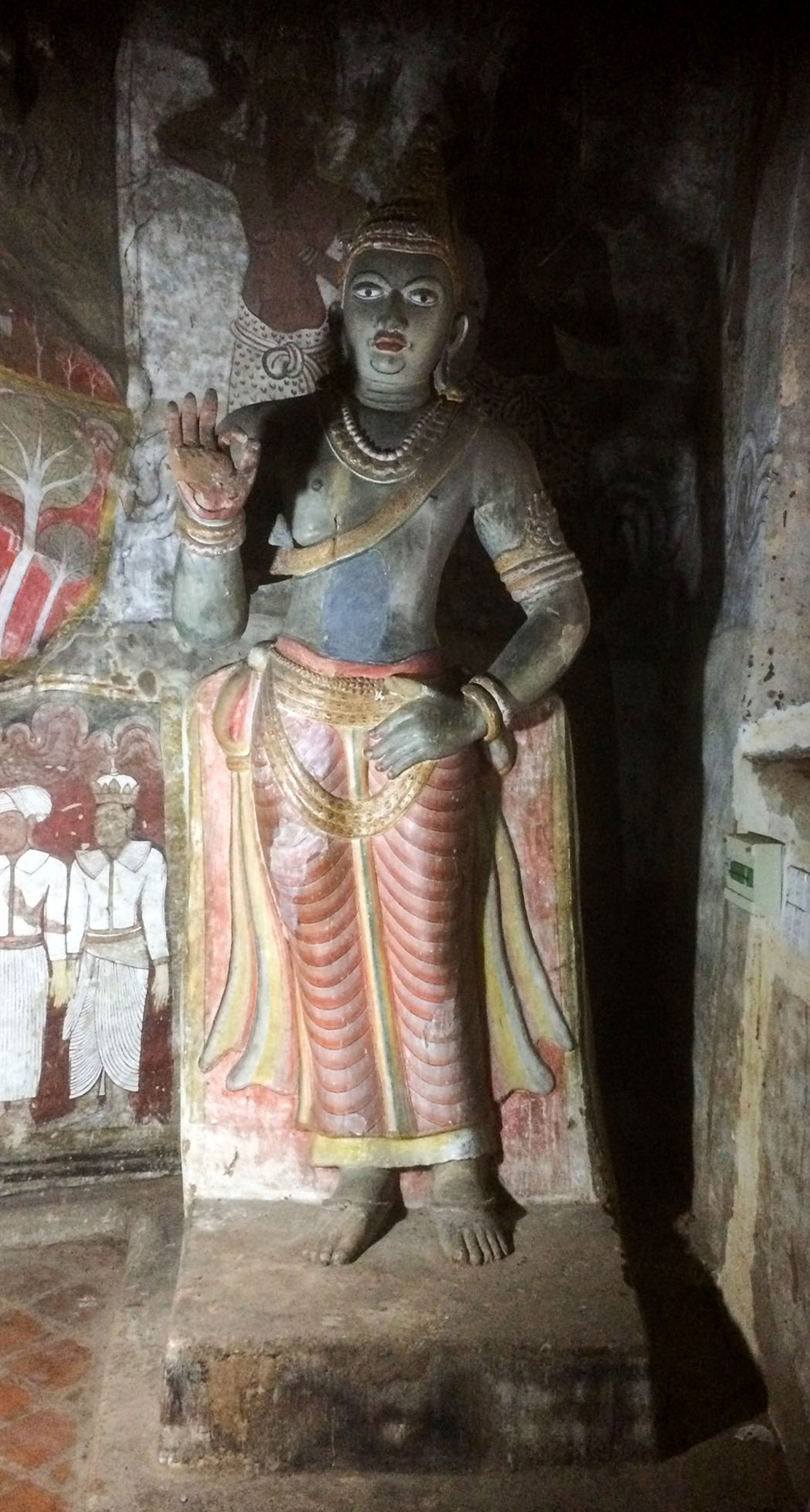 Photograph of a blue coloured statue of Nissanka Malla with the right hand resting on its hip and the other raised in the vitarka mudra inside the Dambulla Caves